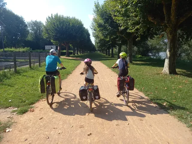 Trois enfants pédalent côte à côte sur une piste cyclable en terrain stabilisé au milieu d'une rangée d'arbres dans l'herbe tondue. Leurs vélos sont chargés avec des sacoches.